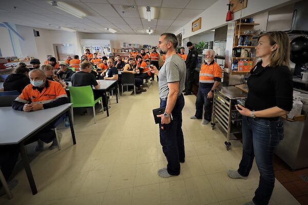 The station leader standing before a room full of station members seated at tables. He is speaking to them about the situation.