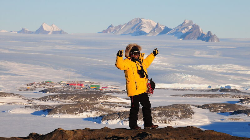 Person wearing yellow cold-weather gear raises their arms with station in the distant background