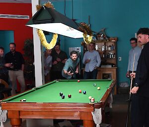 Two people playing a game of billiards. An additional number of people are standing around the billiards table to watch the game. One of the players is lining up his cue to take a shot.