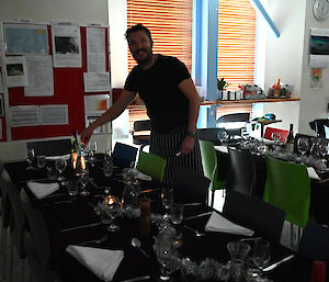 A dining table covered with black cloth and laid out with cutlery, glassware and decorations. One of the mess staff smiles as he lights a candle on the table.