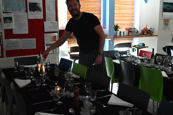 A dining table covered with black cloth and laid out with cutlery, glassware and decorations. One of the mess staff smiles as he lights a candle on the table.