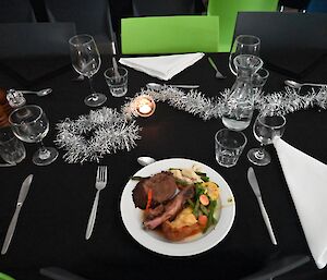 A dinner plate loaded with a selection of meat and vegetables on a dining table. The table is laden with cutlery, glassware, and candles and tinsel for decorations.