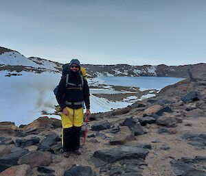 A man wearing yellow and black clothes with a hiking pack standing amongst snow and rocky ground