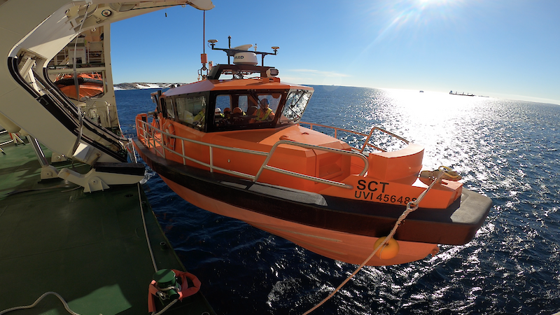 An orange boat with crew inside being launched by davit crane from an icebreaker ship in Antarctica. Calm waters, blue skies, and the Antarctic continent in the background.