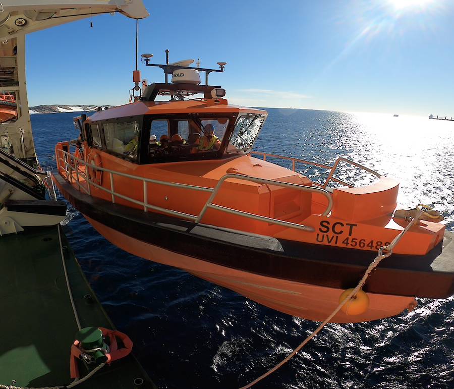 An orange boat with crew inside being launched by davit crane from an icebreaker ship in Antarctica. Calm waters, blue skies, and the Antarctic continent in the background.