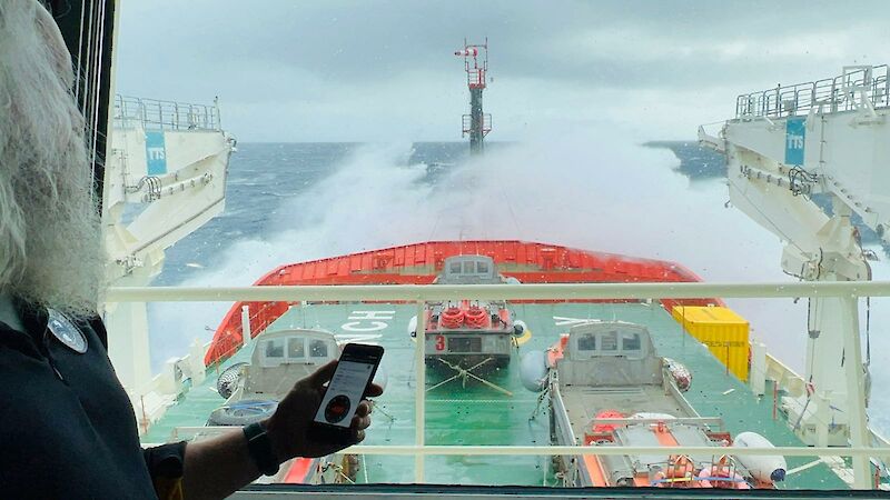 A person holding a mobile device displaying data. Person overlooking forward hatches of icebreaker storing LARCs and wave breaking over bow.