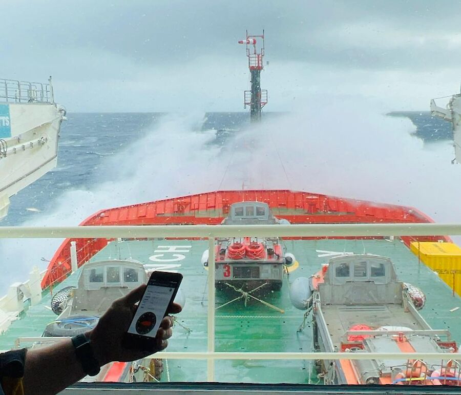 A person holding a mobile device displaying data. Person overlooking forward hatches of icebreaker storing LARCs and wave breaking over bow.