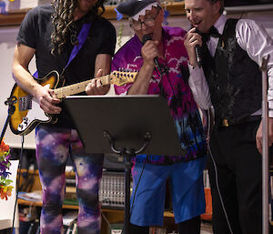 Three people in colourful fancy dress sing karaoke