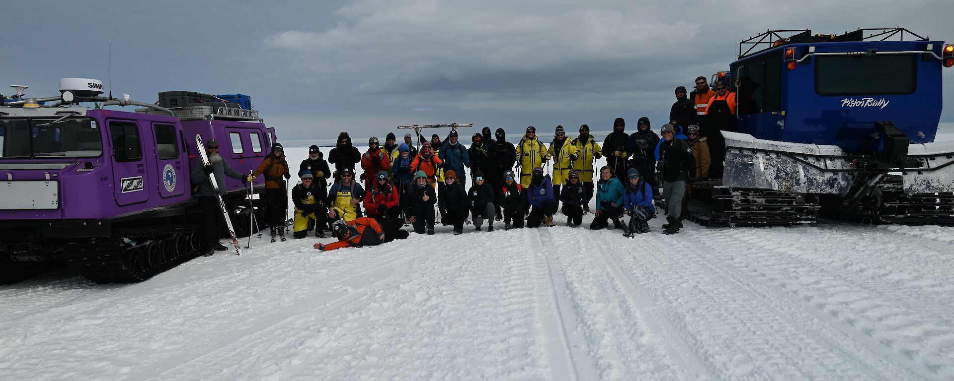 A group of people assembled in two rows for a photo between two snow vehicles. The front row are kneeling, most in the back row are holding skis and ski poles.