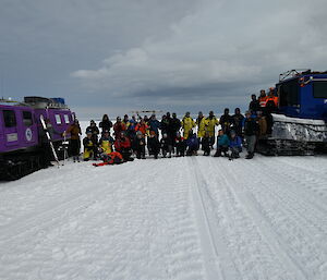 A group of people assembled in two rows for a photo between two snow vehicles. The front row are kneeling, most in the back row are holding skis and ski poles.