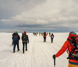 A view from behind of people walking and skiing along a groomed track across a snowy plain, with multiple layers of clouds in the sky above them.