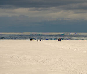 A long distance view of a group of walkers and skiers on the track, and a couple of vehicles travelling with them to the right of the track. In the distance is a view of the ocean, crowded with floating ice and a small iceberg.
