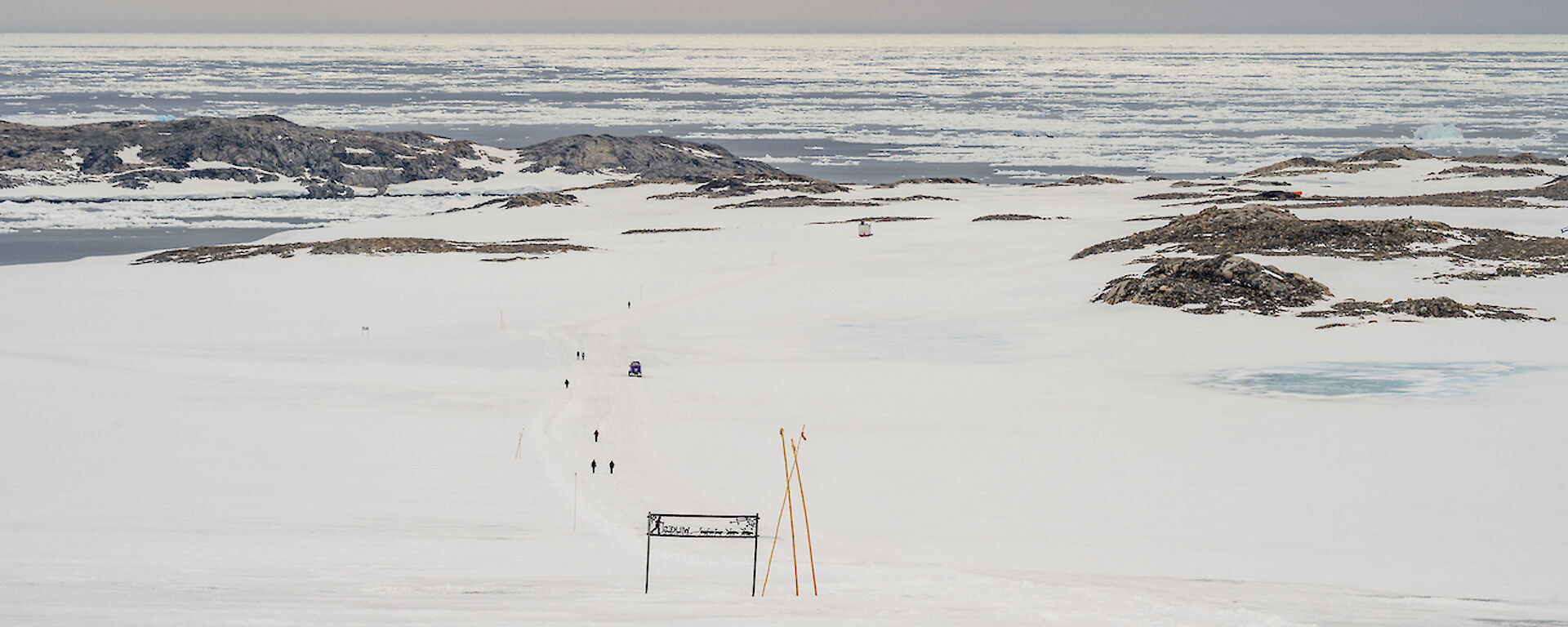 A long distance view of walkers on a curving part of the track going downhill. In the middle ground are low, rocky hills, against an ice-filled ocean background.