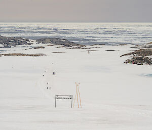 A long distance view of walkers on a curving part of the track going downhill. In the middle ground are low, rocky hills, against an ice-filled ocean background.