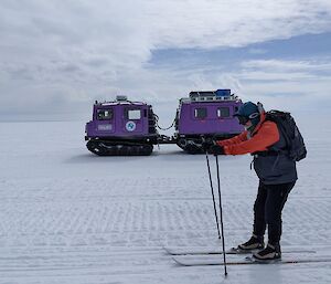 In the foreground, a skier is making his way down the track. In the background, to the skier's right, is a purple Hägglunds vehicle on treads