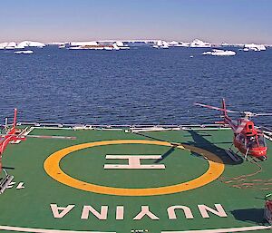2 red helicopters on helideck of icebreaker in Antarctica, ice bergs and rocky islands, calm waters and blue skies.