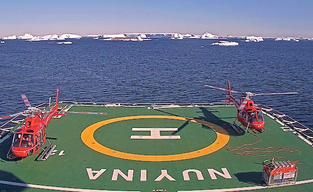 2 red helicopters on helideck of icebreaker in Antarctica, ice bergs and rocky islands, calm waters and blue skies.