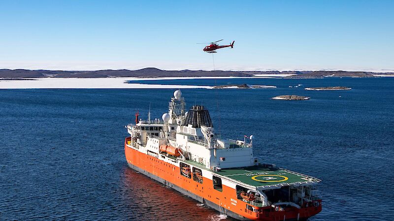 An aerial drone photo of an orange and white ship sailing through the ocean. A red helicopter hovers above the ship.