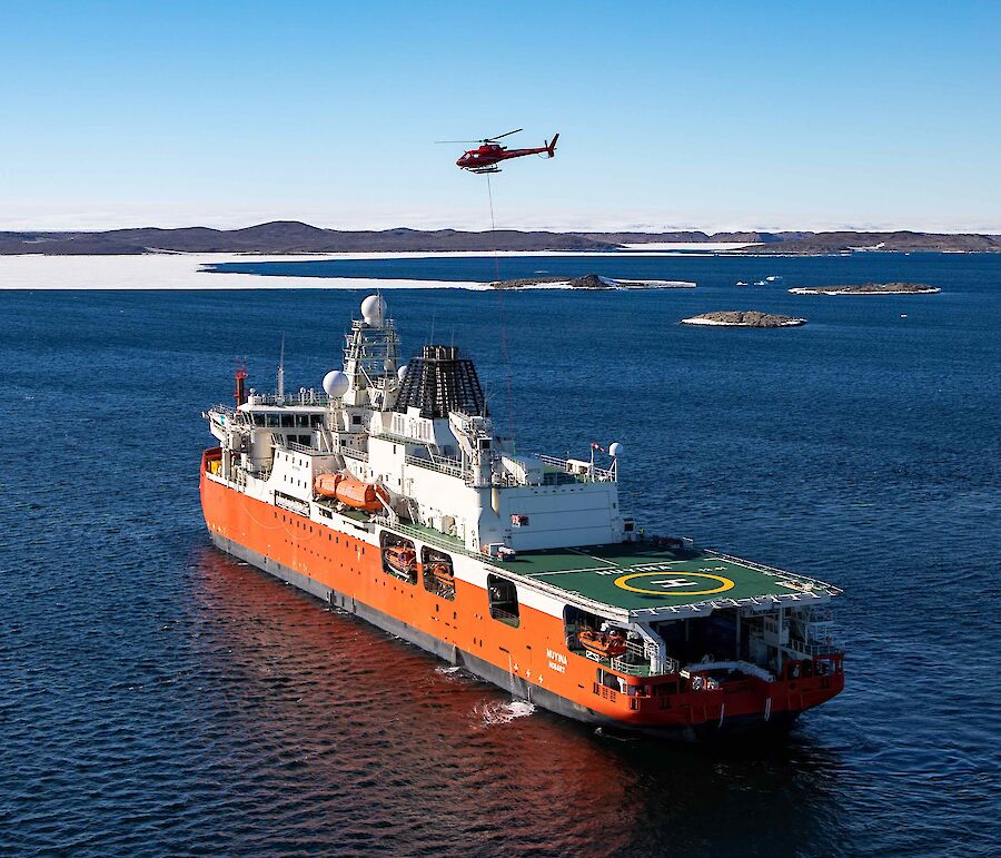 An aerial drone photo of an orange and white ship sailing through the ocean. A red helicopter hovers above the ship.