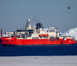 An orange and white ship with ice formations in the background. A red helicopter flies above, carrying cargo from the ship.