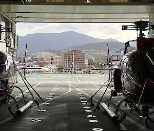 Inside the cargo hold of a ship, 2 helicopters are parked side by side. Hobart city is visible through the open roller door.