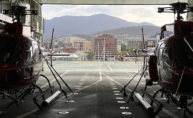 Inside the cargo hold of a ship, 2 helicopters are parked side by side. Hobart city is visible through the open roller door.