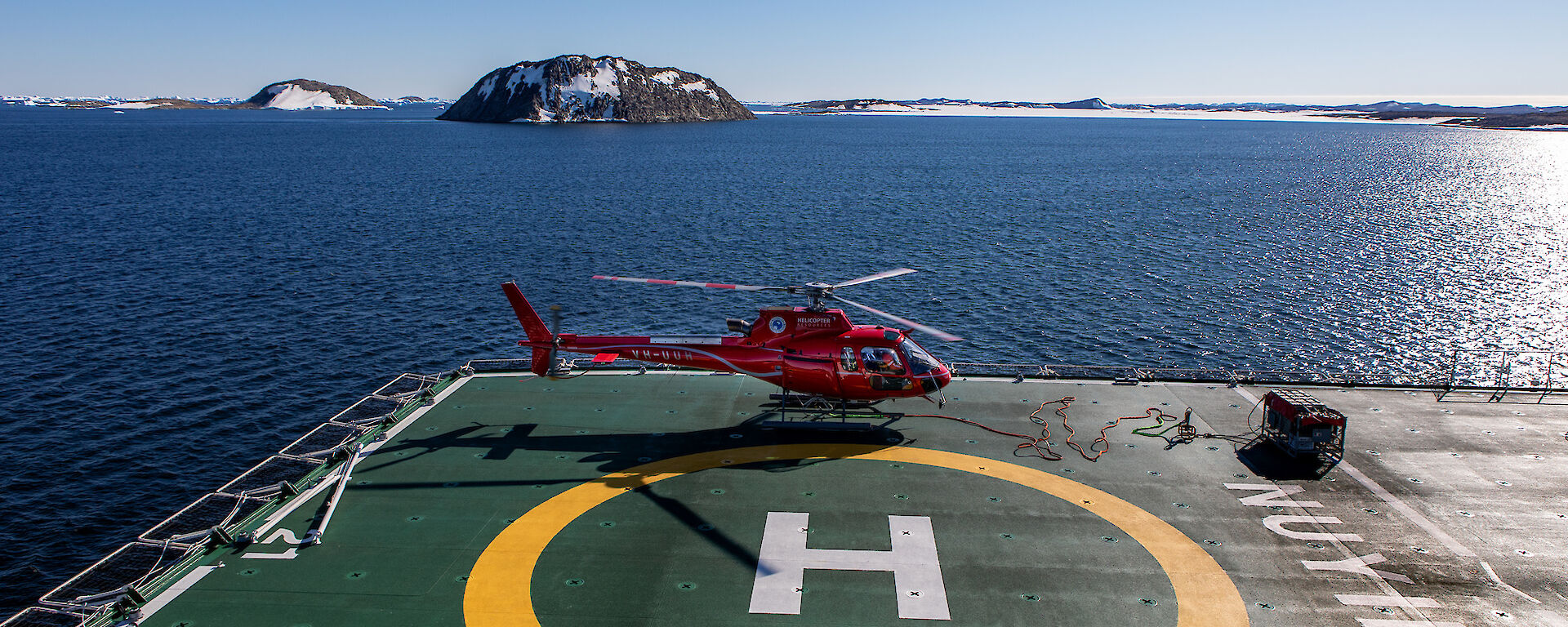 A red helicopter sits on the deck of a ship. In the background is blue water and a number of islands with snow.