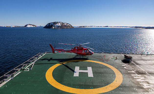 A red helicopter sits on the deck of a ship. In the background is blue water and a number of islands with snow.