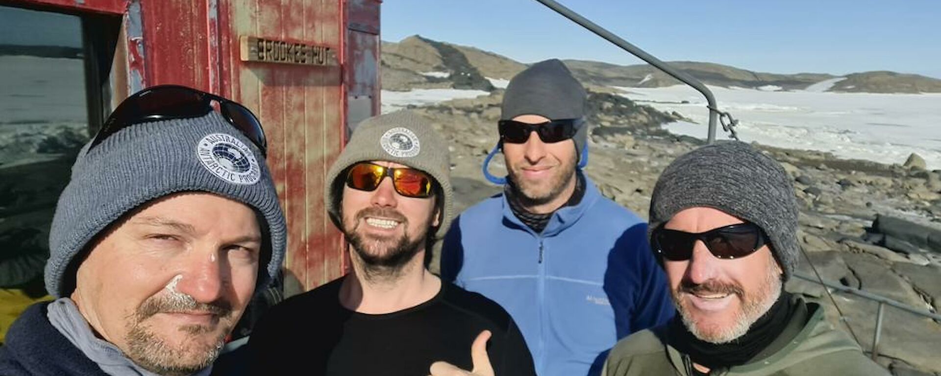 4 men in beanies standing outside a faded red hut