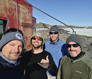 4 men in beanies standing outside a faded red hut