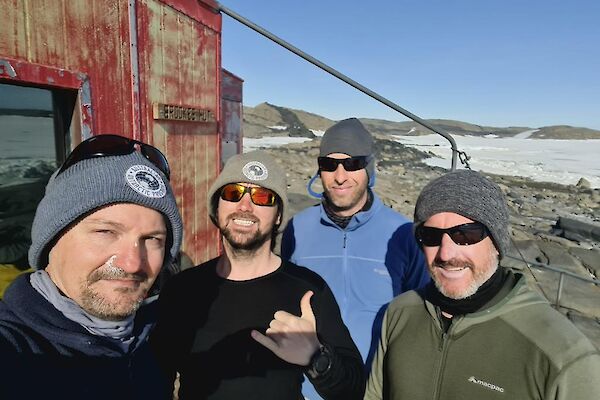4 men in beanies standing outside a faded red hut