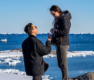 A young couple standing on top of a hill overlooking the ocean. The man is proposing marriage to the woman, offering her an engagement ring