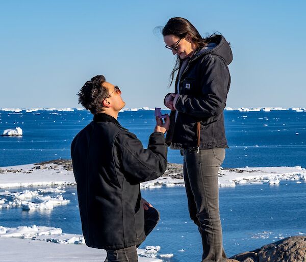 A young couple standing on top of a hill overlooking the ocean. The man is proposing marriage to the woman, offering her an engagement ring
