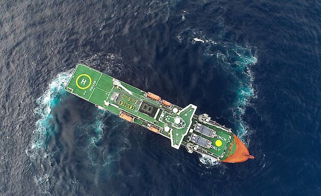 An aerial drone photo. Aerial view of an icebreaker ship in open water. Trails of bubbles and light blue water show how the ship is turning in the water.