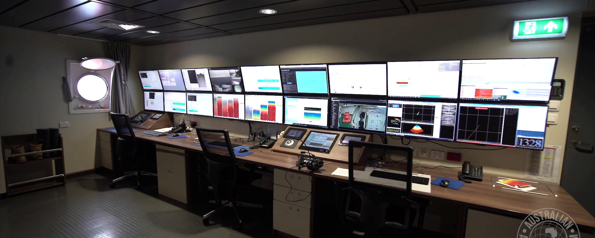 A long desk with control panels and chairs in front of a wall of 18 screens in a room on the ship with a ship portal window.