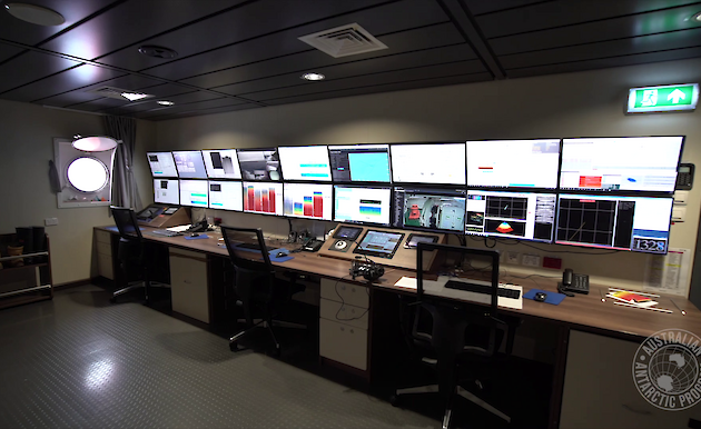 A long desk with control panels and chairs in front of a wall of 18 screens in a room on the ship with a ship portal window.