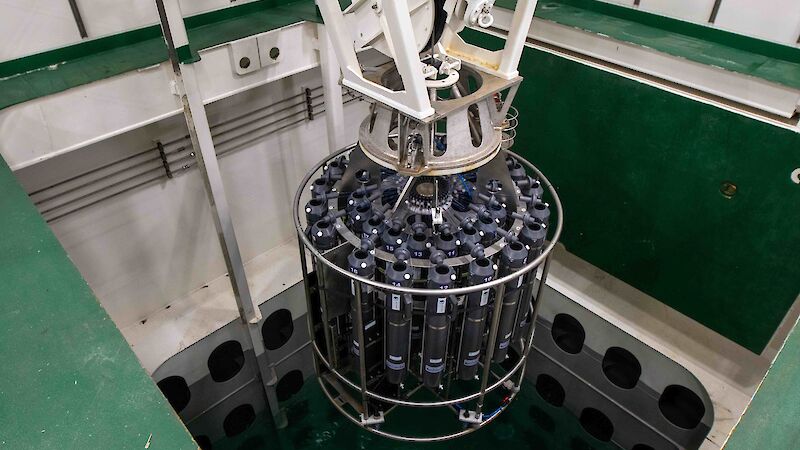 A rosette of water sampling bottles in a round steel frame is lowered by a crane in to a pool of water in the ship's hull