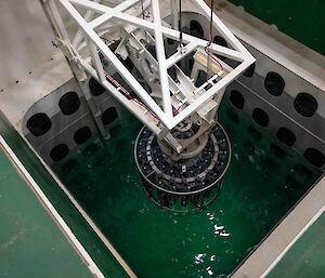 A rosette of water sampling bottles in a round steel frame is lowered by a crane in to a pool of water in the ship's hull