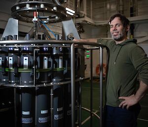 man standing beside a rosette of large water sampling bottles inside a ship