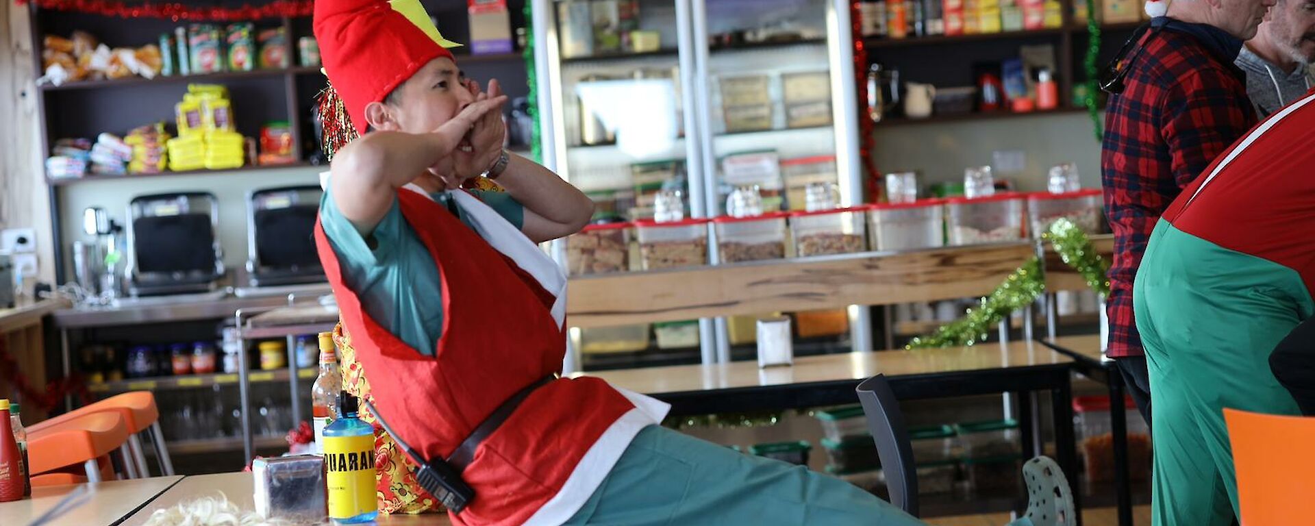 A man dressed as Santa's elf sitting on the edge of a table yelling instructions to a team