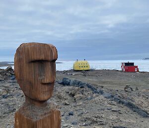 A wooden carved statue outside on rocky ground with some small building structures in the background