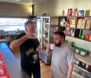 Two men sipping out of mugs standing in the mess area