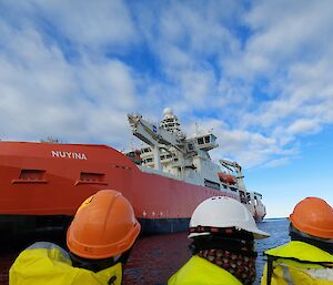 A view of the ship RSV Nuyina from a boat as it approaches the ship. Three passengers on the boat, wearing hard hats and Gore-Tex jackets, are looking up at the ship