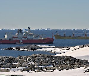 Two large ships, one with a red hull, the other with a yellow hull, beside each other in the ocean, viewed over a rocky and snowy coastline. A number of icebergs are floating in the background, on the ocean horizon