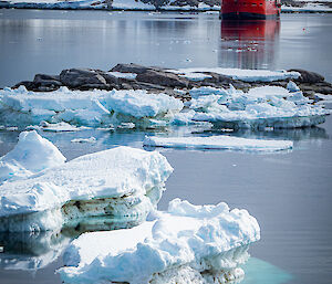 The ship RSV Nuyina resting on calm water in the bay, seen over a rocky islet and a few bits of sea ice