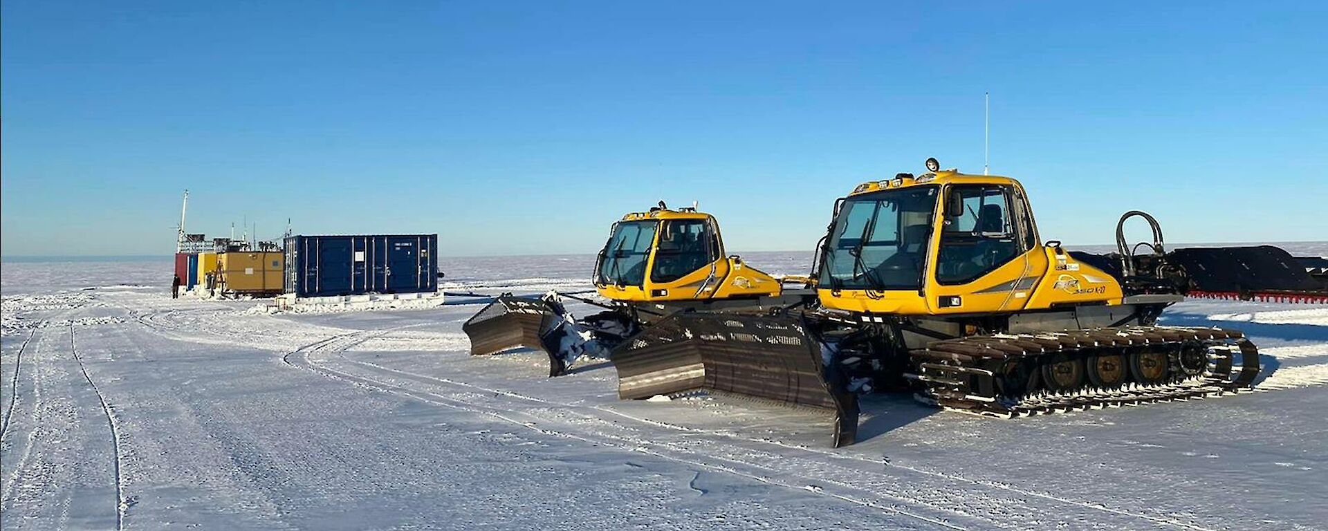 A couple of yellow snow groomers with some shipping containers in the background all sitting on the ice plateau