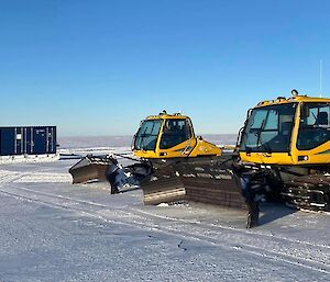 A couple of yellow snow groomers with some shipping containers in the background all sitting on the ice plateau