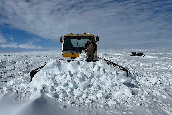 A man stands on the snow in front of a snow groomer moving snow at Woop Woop skiway