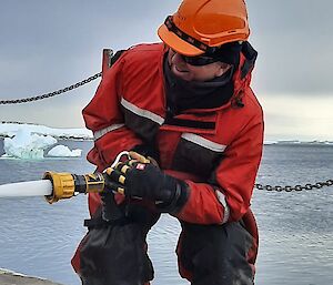 A cargo worker wearing an orange hard hat and a red protective suit, crouching down and discharging a high-powered hose at an object out of frame