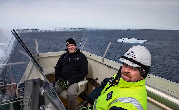 two men on deck of the ship with glacier in background